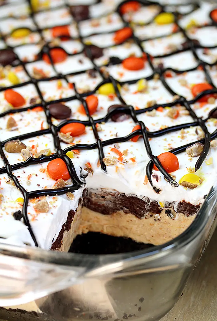 Reese‘s Pieces Peanut Butter Chocolate Lasagna – simple and easy, no bake, layered, rich taste dessert, with Reese‘s pieces, Oreo cookies, peanut butter and chocolate pudding topped with cool whip, Reese‘s pieces, and chocolate syrup.