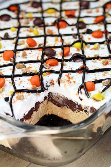 Reese‘s Pieces Peanut Butter Chocolate Lasagna – simple and easy, no bake, layered, rich taste dessert, with Reese‘s pieces, Oreo cookies, peanut butter and chocolate pudding topped with cool whip, Reese‘s pieces, and chocolate syrup.