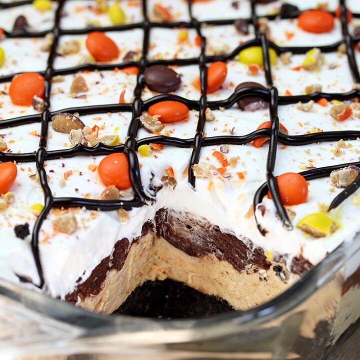 Reese‘s Pieces Peanut Butter Chocolate Lasagna – simple and easy, no bake, layered, rich taste dessert, with Reese‘s pieces, Oreo cookies, peanut butter and chocolate pudding topped with cool whip, Reese‘s pieces, and chocolate syrup.