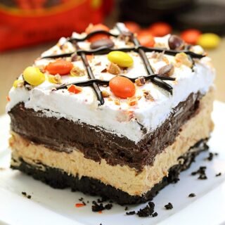 Reese‘s Pieces Peanut Butter Chocolate Lasagna – simple and easy, no bake, layered, rich taste dessert, with Reese‘s pieces, Oreo cookies, peanut butter and chocolate pudding topped with cool whip, Reese‘s pieces, and chocolate syrup.
