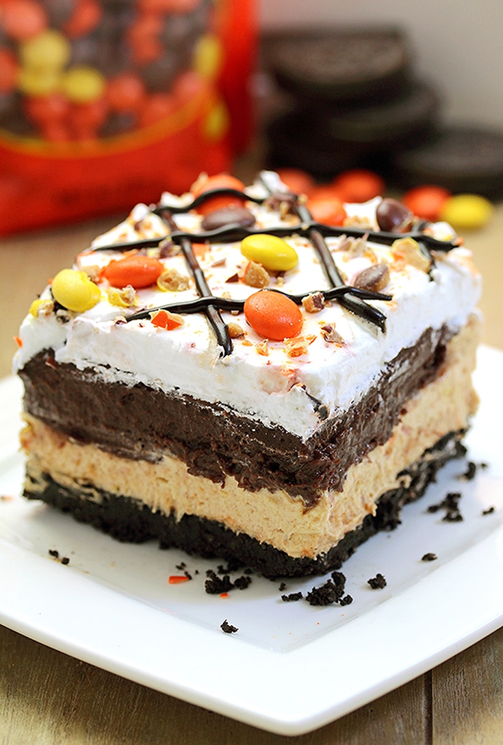 Reese‘s Pieces Peanut Butter Chocolate Lasagna – simple and easy, no bake, layered, rich taste dessert, with Reese‘s pieces, Oreo cookies, peanut butter and chocolate pudding topped with cool whip, Reese‘s pieces, and chocolate syrup.