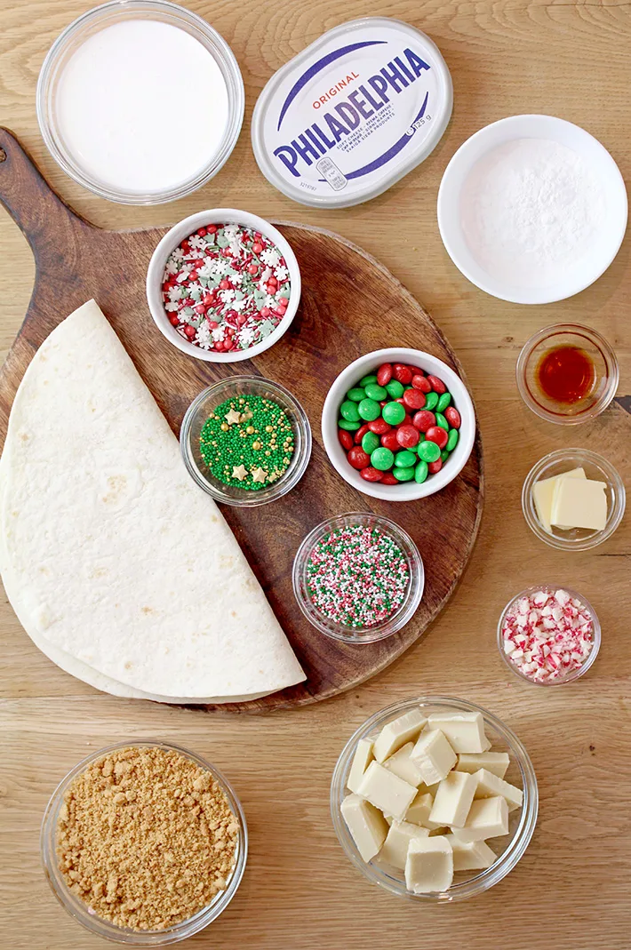 Let’s start with the ingredients that are necessary to prepare these festive and delicious treats:Flour tortillas, graham cracker crumbs, unsalted  butter, white chocolate, cream cheese, powdered sugar, vanilla extract, heavy whipping cream, Christmas sprinkles, M&M's, candy canes.