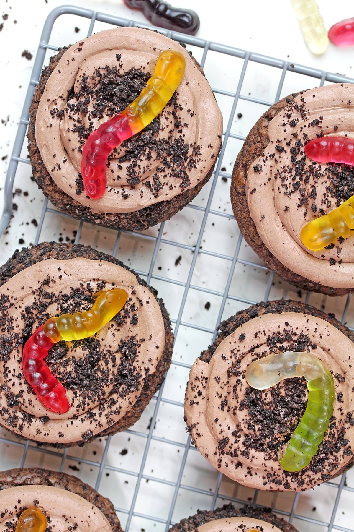 Chocolate cookies topped with chocolate mousse, Oreo crumbs, and gummy worms on a wire rack.